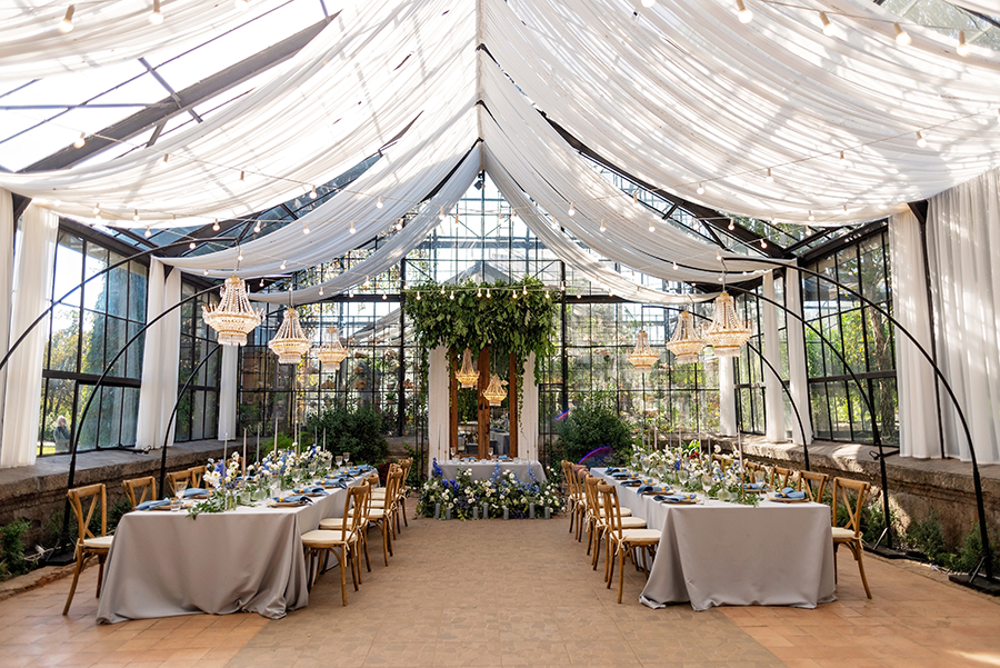 Elegant event setup inside a glass greenhouse with a draped fabric ceiling, chandeliers, and two long dining tables decorated with flowers and place settings.