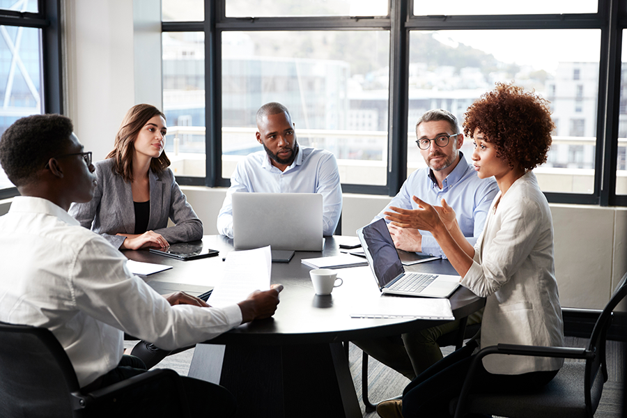 Group of five colleagues sitting around a conference table with laptops and papers, engaged in a team discussion.