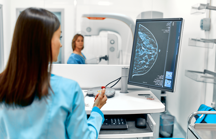 A technician stands at computer equipment with a screen displaying a mammogram.