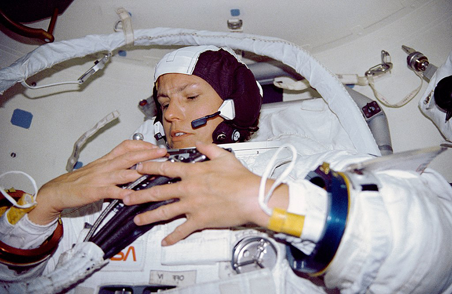 Astronaut inside a spacecraft wearing a spacesuit and communication headset, adjusting equipment with both hands.