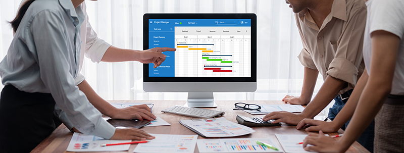 A group of people stand around a table reviewing printed charts while looking at a computer monitor displaying a project schedule with color-coded timeline bars.