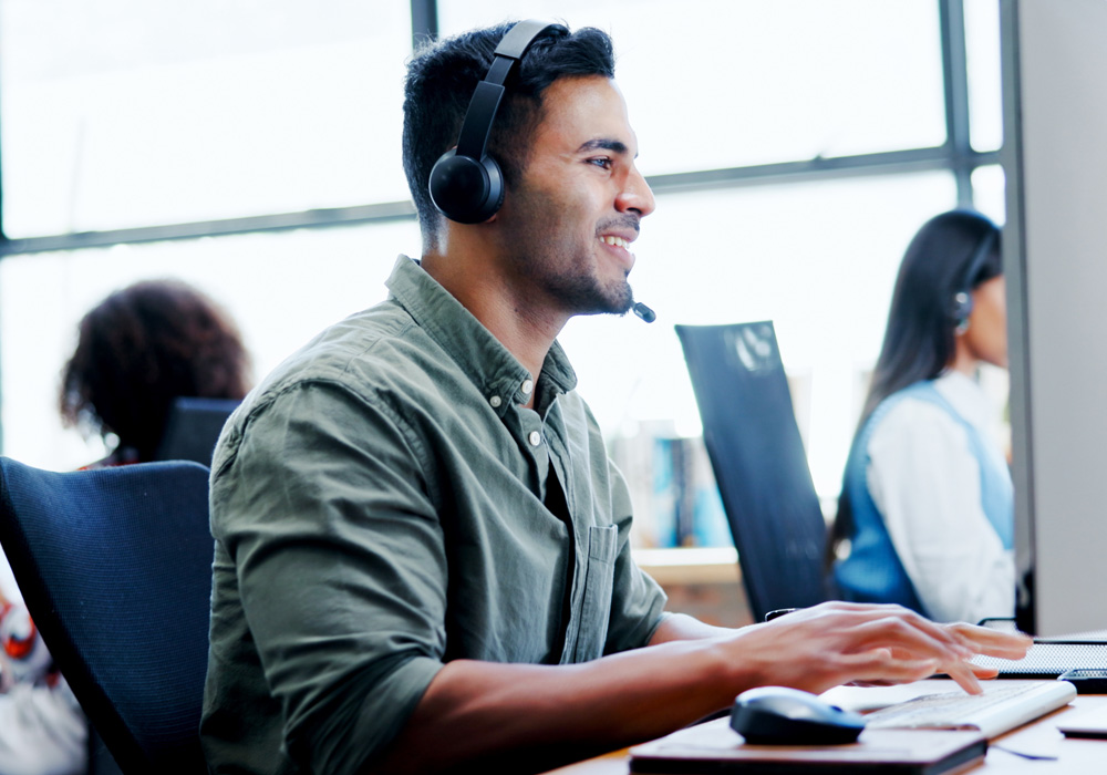 Man wearing a headset smiling and speaking while on a video call, seated at a desk with a laptop and coffee mug in a modern office setting
