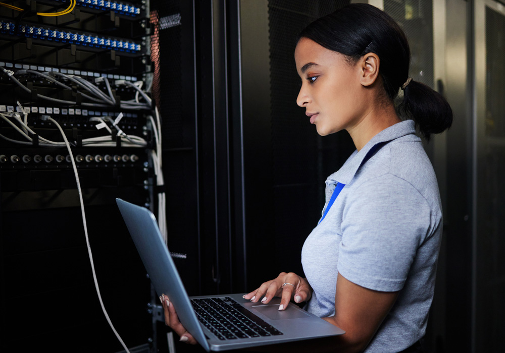 Woman working on a laptop in a server room, standing beside network equipment racks with visible cables and data projections on the screen