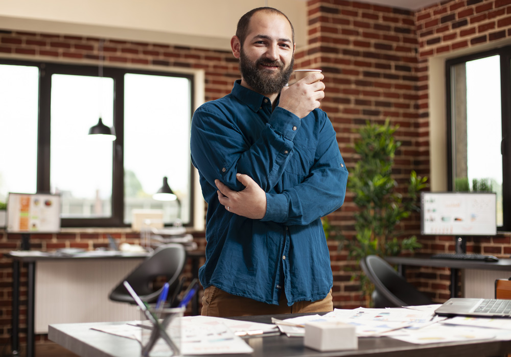 Smiling man standing with arms crossed in a bright office, with a laptop, papers, and plants visible on the desk behind him