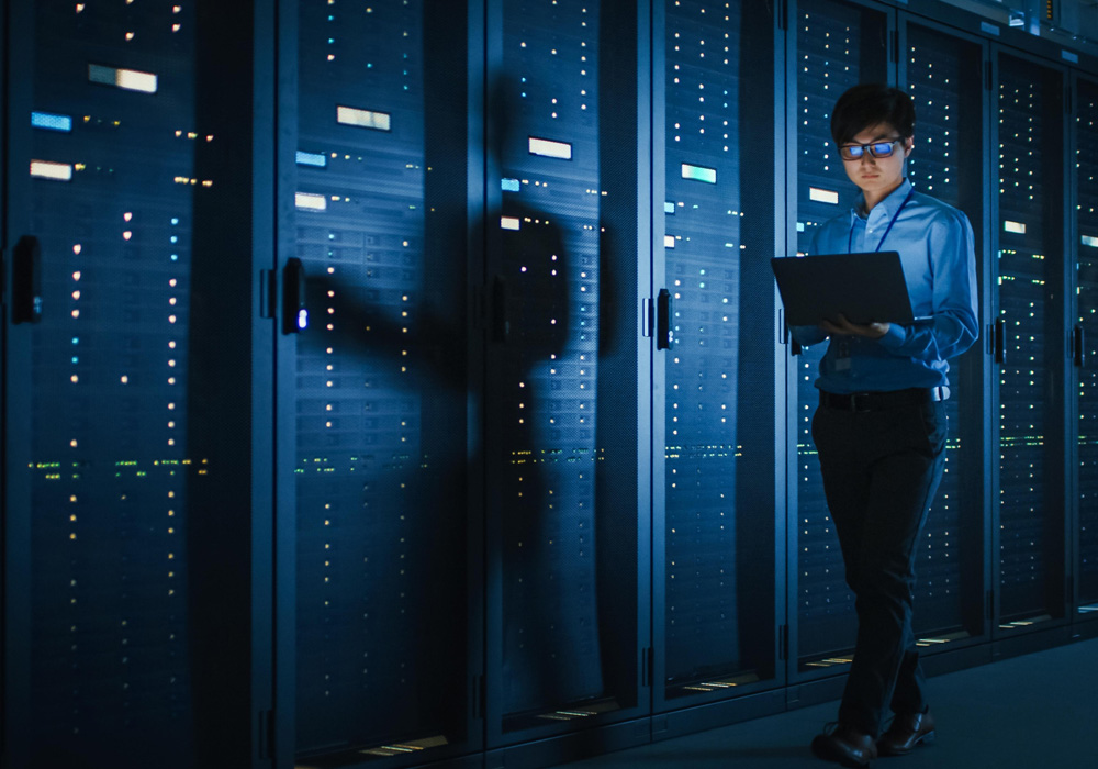 IT professional walking through a data center aisle lined with illuminated server racks, holding a laptop to monitor systems