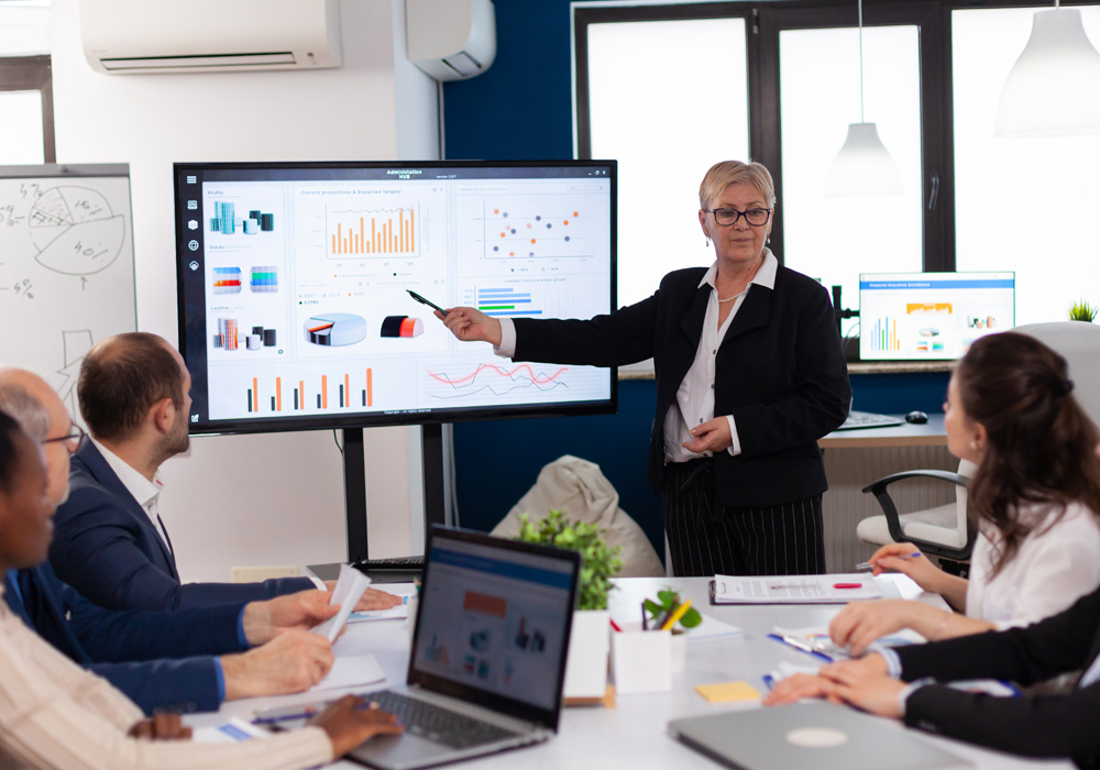 Team in a modern office conference room watching a presenter point to a large screen displaying data charts and performance graphs for networks