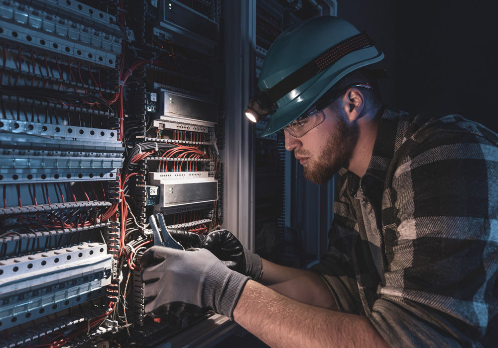 Technician wearing a grey cap and headlamp, inspecting or repairing complex wiring in a telecommunications or network server panel