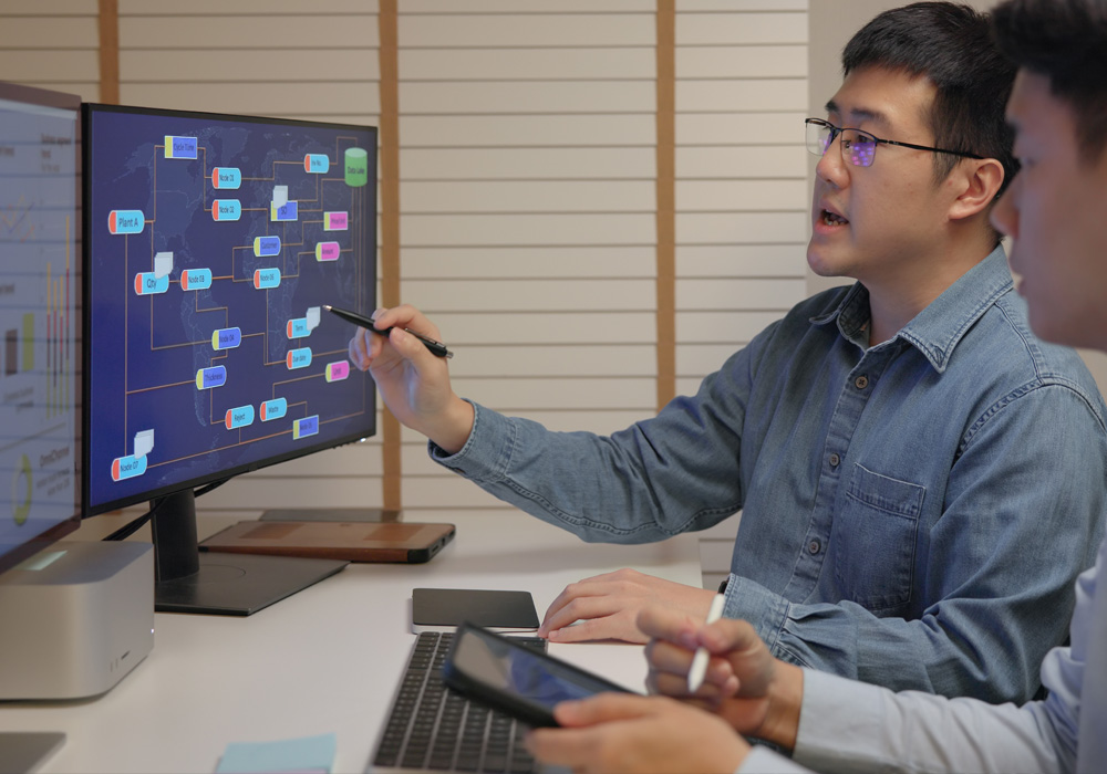Two men collaborating at a desk with dual monitors, discussing a colorful flowchart or data diagram displayed on the screen