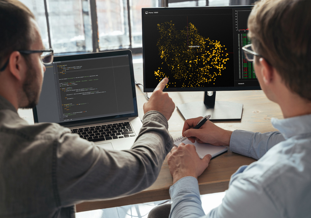  Two men analyzing data on computer screens, with one showing code and the other displaying a visualization of yellow data points on a black background